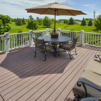 Eye-level view of a wooden deck with light-brown deck chairs, a table with an umbrella and white railings surrounding its border. There are flowers on the table and a plant pot with a shrub next to the chairs. There is green grass, trees and homes in the background.
