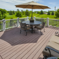Eye-level view of a wooden deck with light-brown deck chairs, a table with an umbrella and white railings surrounding its border. There are flowers on the table and a plant pot with a shrub next to the chairs. There is green grass, trees and homes in the background.
