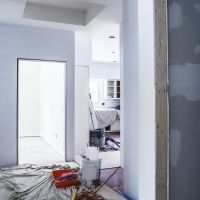 Painting and other construction equipment, tools and materials are scattered on the floor of this view looking through the hallway of an incomplete apartment home addition construction project with high, open ceilings. A plaster patched wall bathroom with a fully framed, unfinished doorway is in the immediate foreground. A brightly lit bedroom is on the far left. and the kitchen opens up around the far right into the rest of the in-law apartment.