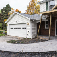 A brand new freshly poured and stamped concrete walkway ramp at a residential home addition in-law apartment construction site. Temporary vertical wooden beams are supporting the new front porch roof in proper position during the building process. The new walkway is designed as a ramp for easy handicap access to the apartment addition entrance door located off camera to the right of the current house front door visible under the new porch roof.